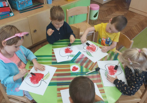 Dzieci siedzą przy stoliku i kolorują obrazek przedstawiający jabłko