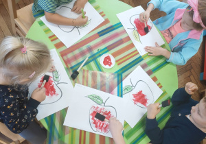 Dzieci siedzą przy stoliku i kolorują obrazek przedstawiający jabłko