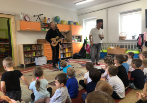 Pan gra na gitarze, drugi pan śpiewa. Dzieci siedzą na dywanie