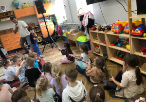 Wokalista z chłopcem trzymają linę, z drugiej strony cztery dziewczynki siedzą na dywanie, trzymają linę