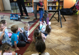 Wokalista z chłopcem trzymają linę, z drugiej strony cztery dziewczynki siedzą na dywanie, trzymają linę