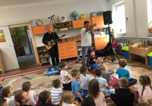 Pan gra na gitarze, drugi pan śpiewa. Dzieci siedzą na dywanie