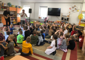 Pan gra na gitarze, drugi pan śpiewa. Dzieci siedzą na dywanie