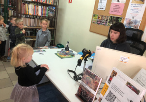 Dziewczyna stoi przy biurku. Pani siedzi za komputerem