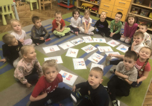 Dzieci siedzą w kole na dywanie. Po środku rozłożone są karty
