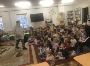 Dzieci siedzą na dywanie. Pani podaje dzieciom instrument