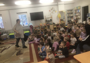 Dzieci siedzą na dywanie. Pani podaje dzieciom instrument